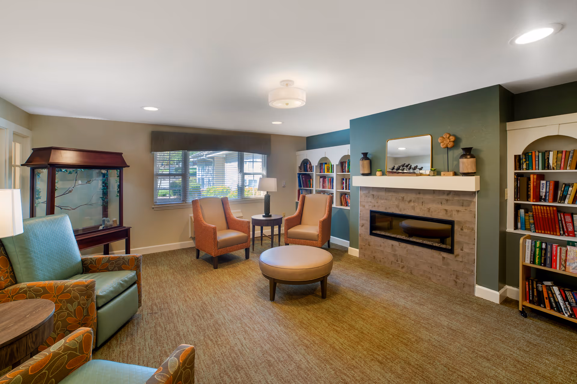 Cozy common living room with upholstered chairs around a round ottoman, a fireplace with a mantel, bookshelves, and a window.
