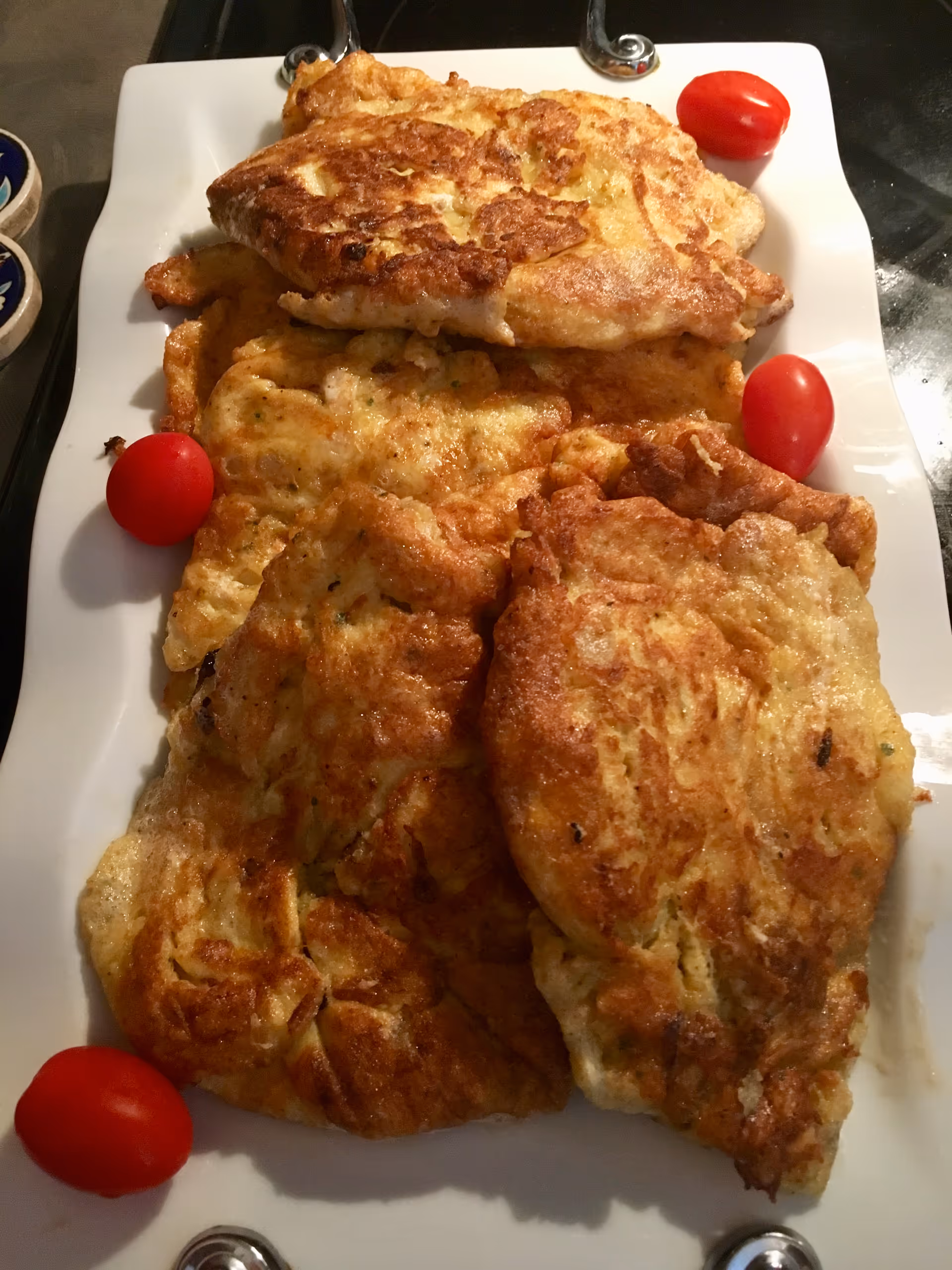 A white rectangular plate with several pieces of golden-brown fried food, possibly fritters or pancakes, garnished with four small red cherry tomatoes placed around the edges.
