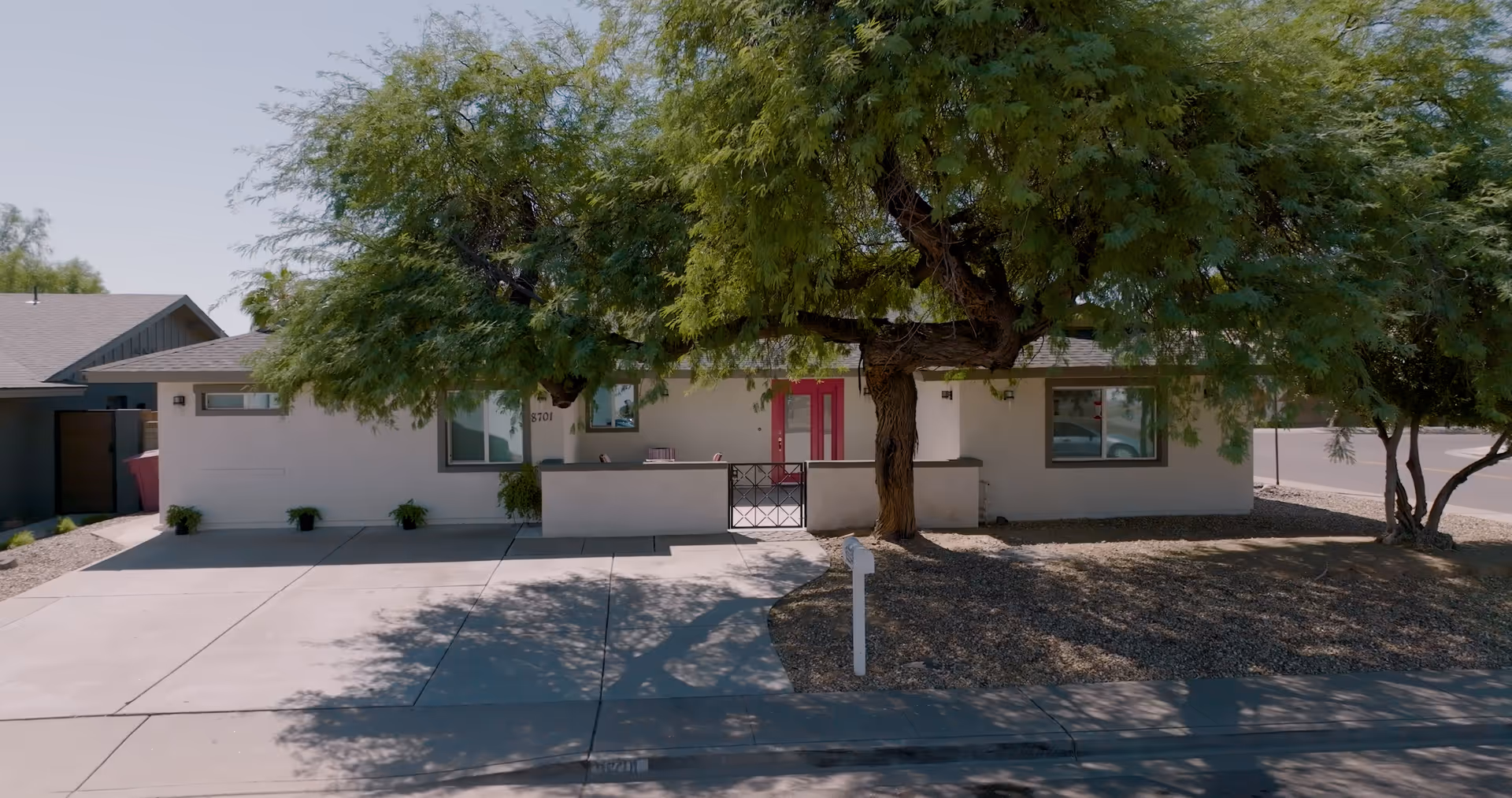 Single-story building front with a large tree shading a driveway, pink double entry doors, and gravel landscaping.