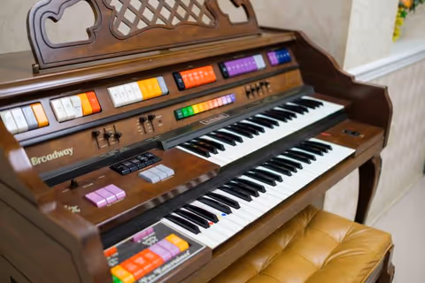 Close-up view of a vintage Broadway electric organ with colorful buttons and two keyboards, placed in a room with light-colored walls and a cushioned bench in front.