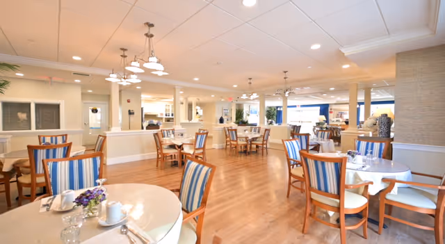 A spacious and well-lit dining room with multiple round tables covered with white tablecloths, each set with cups, glasses, and silverware. The chairs have wooden frames with blue and white striped cushions. The room features wooden flooring, white walls, and ceiling lights, creating a warm and inviting atmosphere.