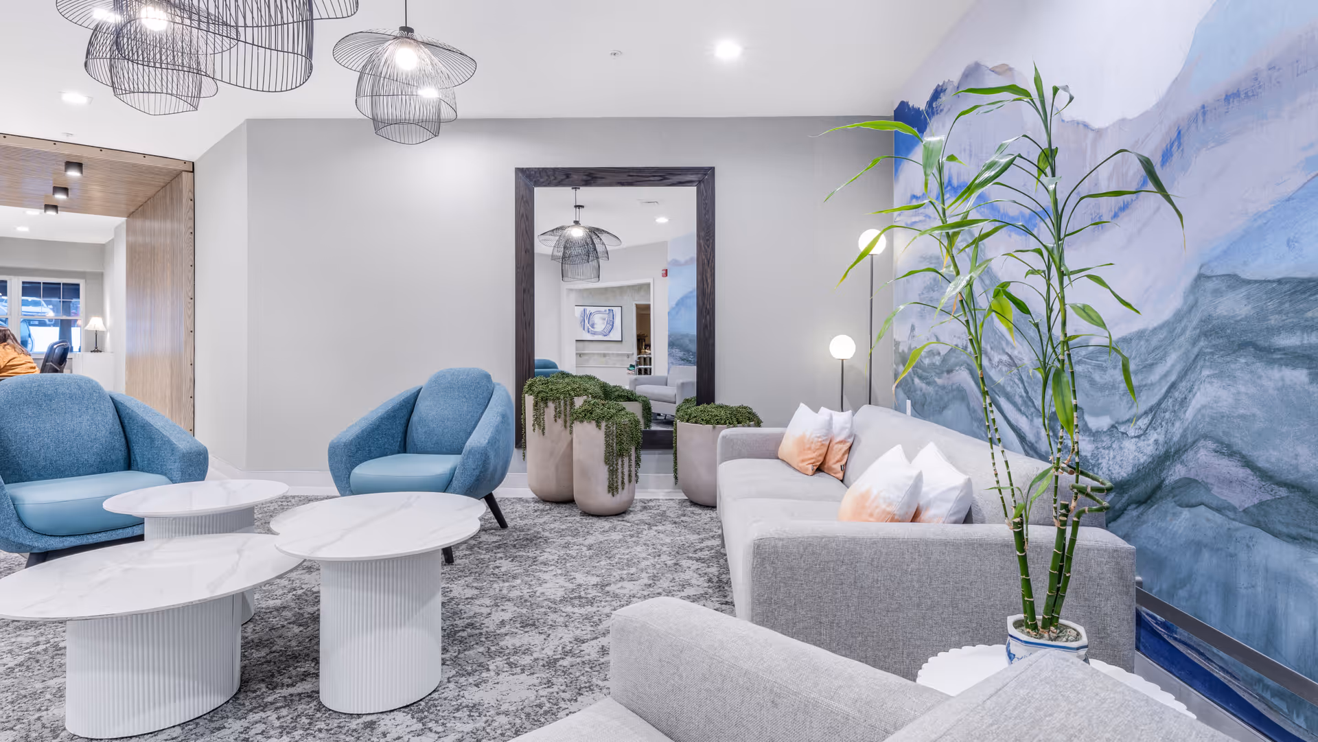 Bright modern lounge with gray sofas, blue armchairs, marble-top coffee tables, potted plants, and a large abstract wall mural.