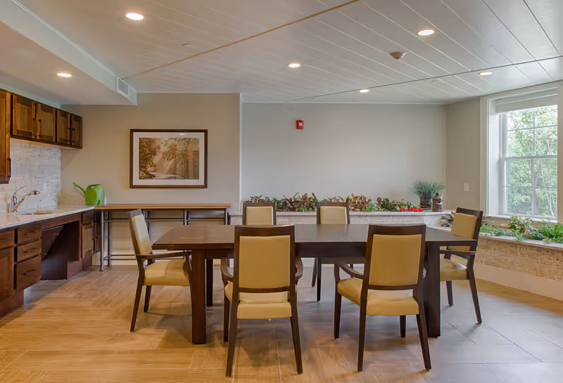 Bright communal dining room with a large wooden table surrounded by upholstered chairs, a kitchenette on the left, and a window with plants on the right.