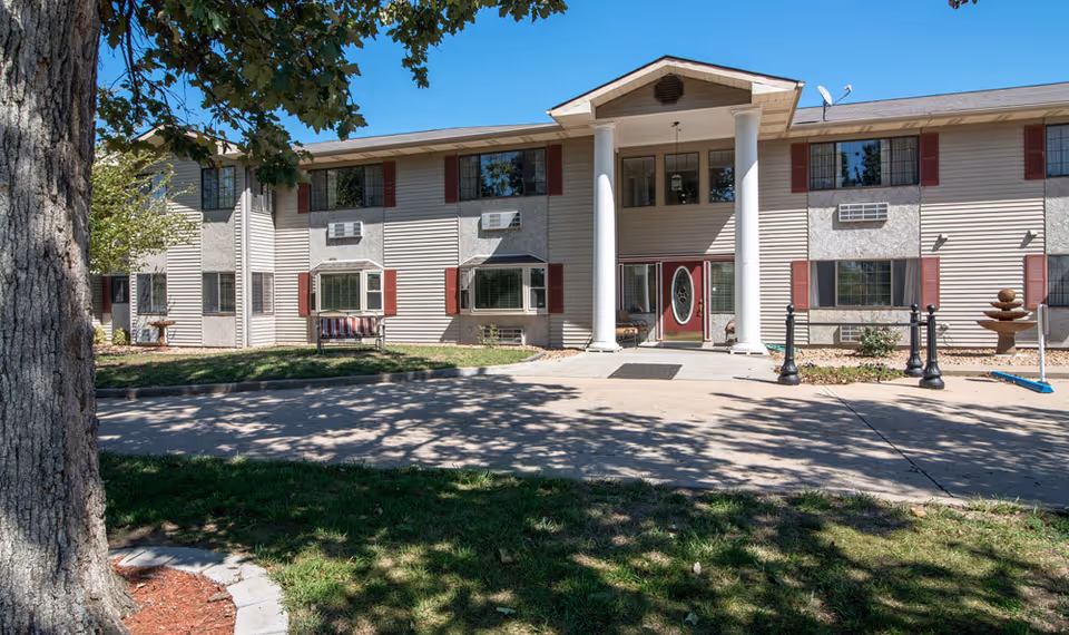 Front exterior view of a two-story senior living facility building with beige siding, red shutters, and white columns at the entrance. There is a concrete driveway and a tree casting shadows on the grass in front of the building.