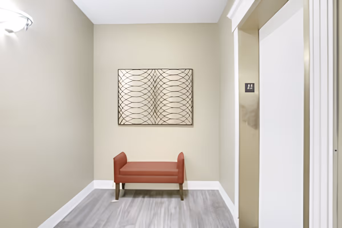 A small hallway or waiting area with a red cushioned bench against a beige wall. Above the bench is a decorative wall art piece with a geometric pattern. To the right, there is an elevator door with a metallic finish and a control panel.