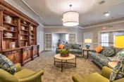 A cozy living room area in a senior living facility featuring patterned sofas with orange and green cushions, two green armchairs, a round wooden coffee table with a plant centerpiece, a large wooden bookshelf filled with decorative items, and a large ceiling light fixture. The room has beige walls and carpet, with glass doors leading to another room in the background.
