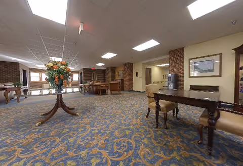Spacious interior common area with patterned carpet, wooden tables and chairs, a large floral arrangement on a round table, brick walls, and framed artwork. The area is well-lit with ceiling lights and has a water dispenser in the background.