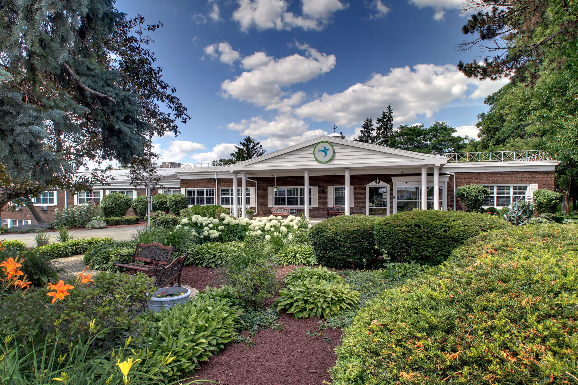 Front entrance of a single-story brick senior living facility with a covered portico, landscaped gardens, and benches.
