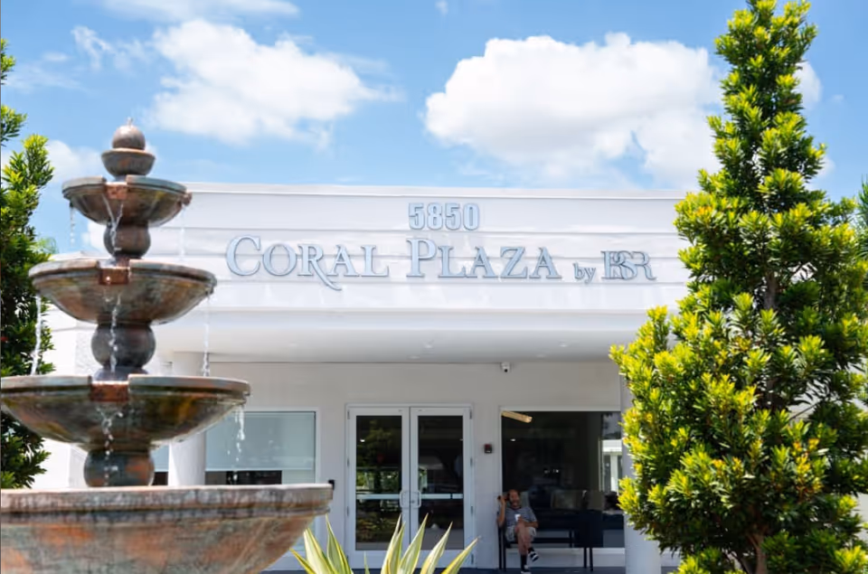 Exterior view of Coral Plaza building entrance with a multi-tiered water fountain in the foreground and green trees on either side under a partly cloudy blue sky.