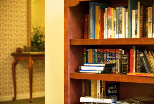 Close-up view of a wooden bookshelf filled with various books in a cozy interior setting. In the background, there is a small wooden table with a decorative plant and a mirror on the wall, partially visible.