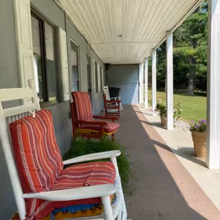 A covered outdoor porch area with white rocking chairs that have red and white striped cushions. The porch has white pillars and overlooks a grassy area with trees and potted plants along the edge.
