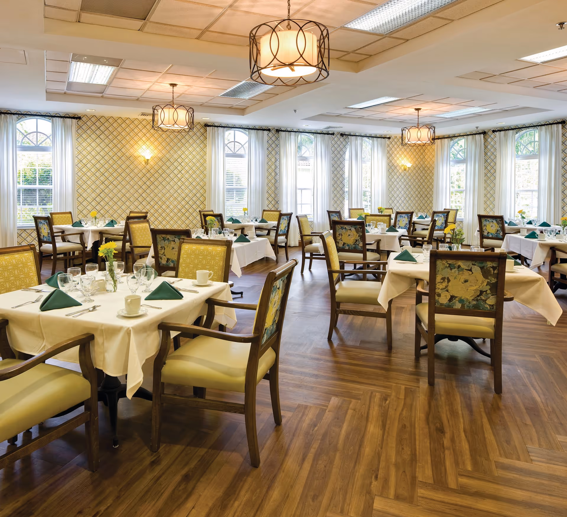 Well-lit dining room with multiple tables set with white tablecloths, green napkins, glassware, and floral-upholstered chairs.
