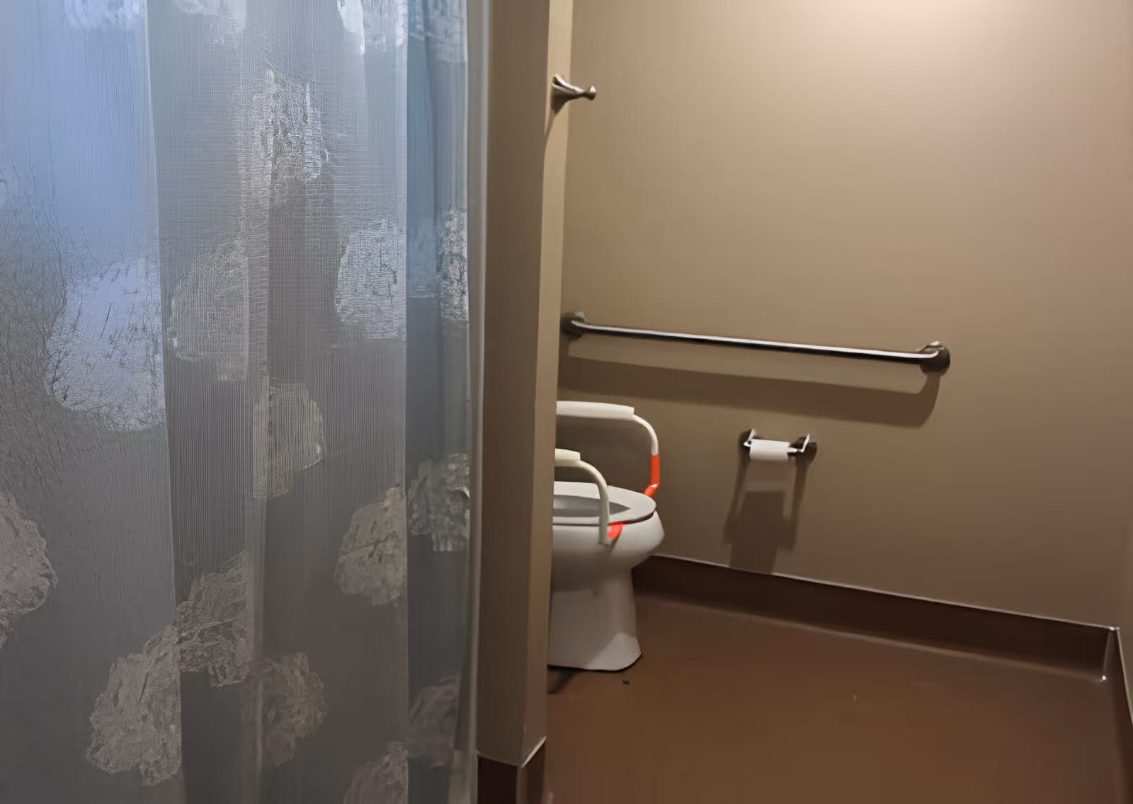 A bathroom with a toilet equipped with white and red safety rails, a grab bar on the wall, a toilet paper holder with a roll of toilet paper, and a shower curtain with a floral pattern partially visible on the left side.
