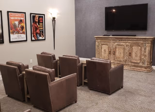 A cozy media room with six brown leather armchairs arranged in two rows facing a large flat-screen TV mounted on a gray wall. Below the TV is an ornate wooden cabinet. The opposite wall features framed classic movie posters and a wall sconce light fixture.