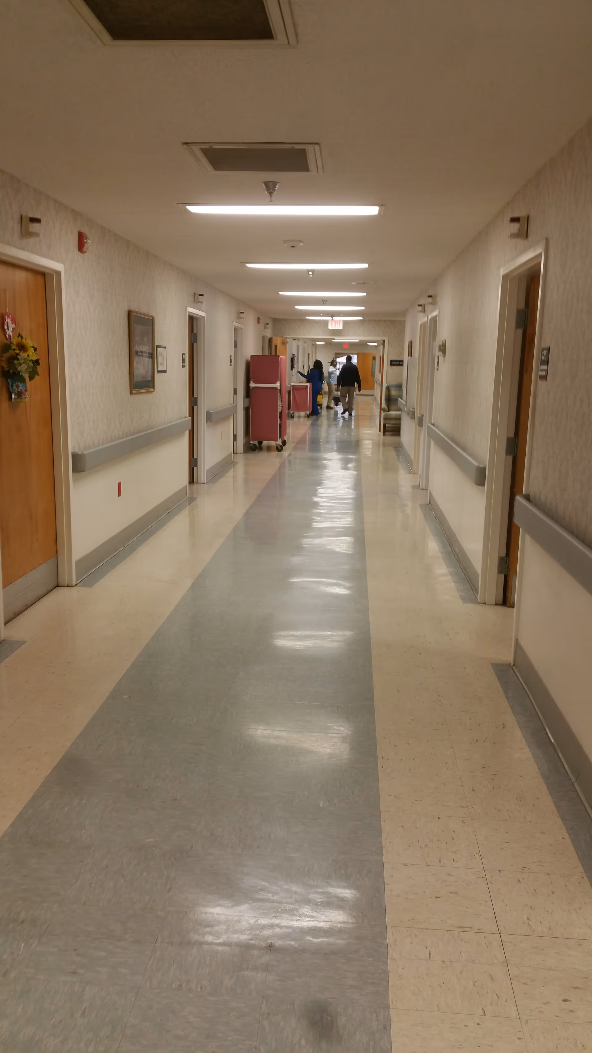 A long, well-lit interior hallway in a senior living facility with linoleum floors, handrails and doors on both sides and staff and carts at the far end.