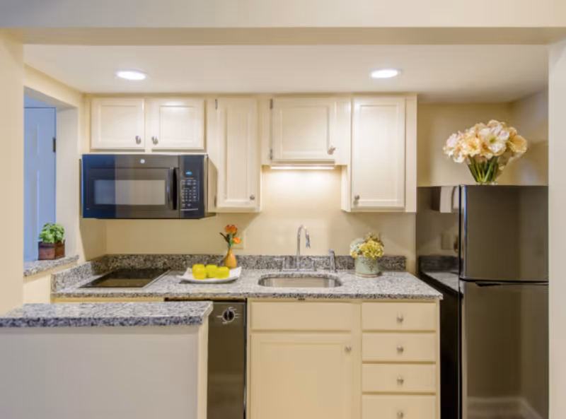 Compact kitchen with granite countertops, sink, microwave, refrigerator, cream cabinets and decorative flowers.