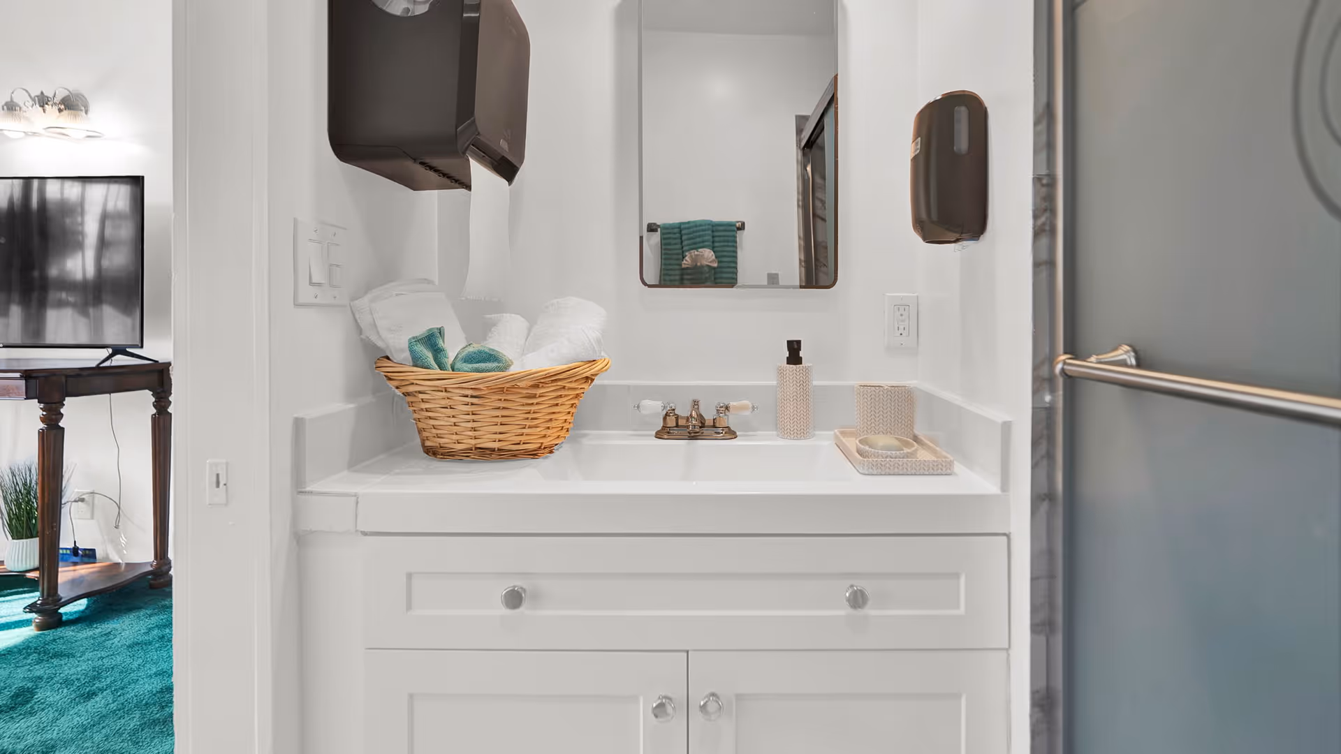 A white bathroom vanity with sink and mirror, a wicker basket of towels, a soap dispenser, and a shower door visible to the right.