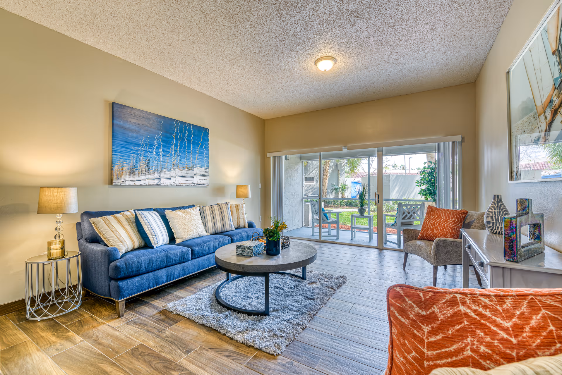 A bright living room with a blue sofa, round coffee table, accent chairs, lamps, and sliding glass doors leading to a patio.