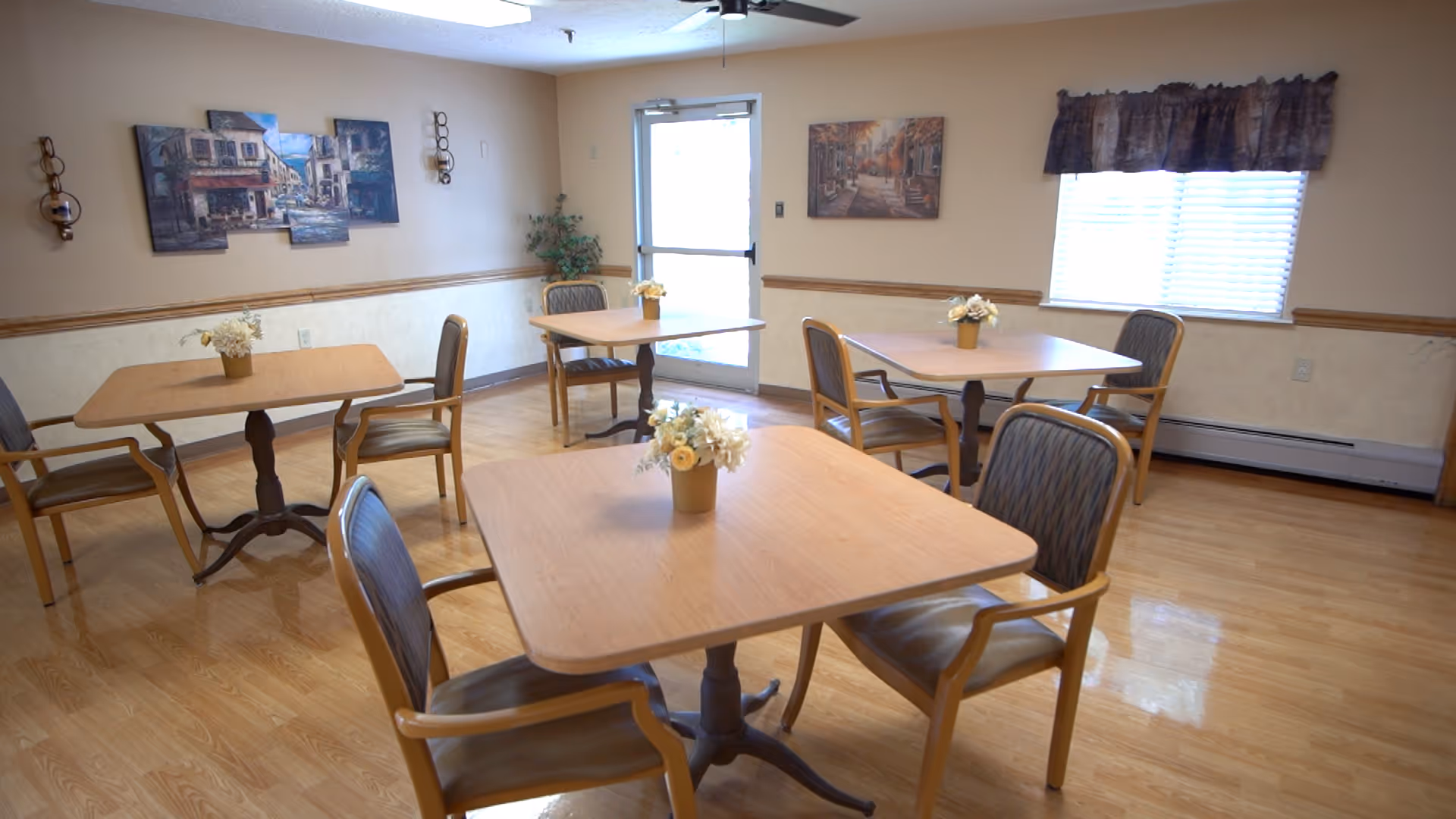 A dining area with four wooden tables, each surrounded by four chairs with cushioned seats and backs. Each table has a small flower arrangement in the center. The room has light-colored walls with wooden trim, a window with a valance, and framed artwork on the walls. The floor is a polished light wood, and there is a door with a window letting in natural light.
