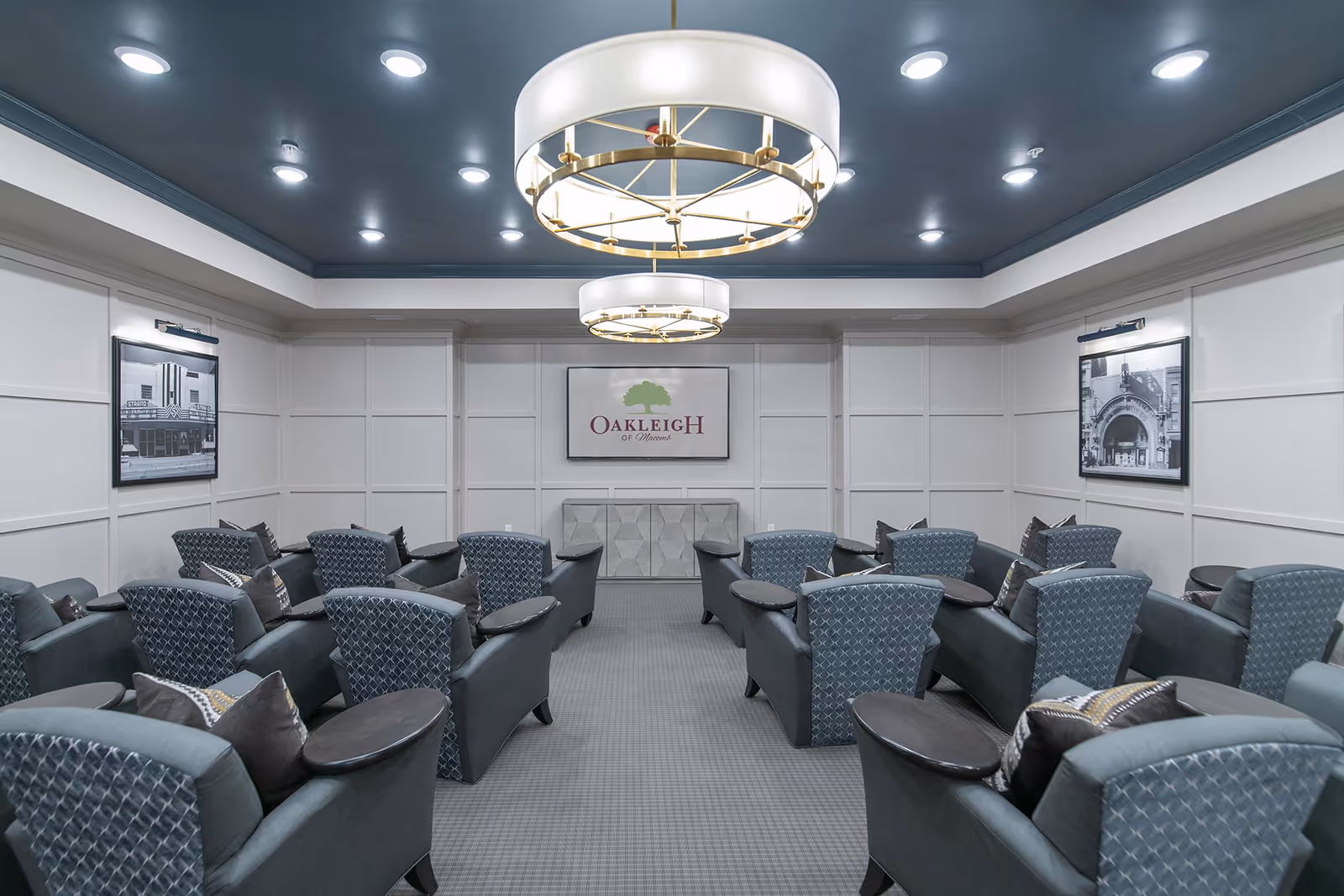 Seating room with rows of upholstered armchairs facing a wall sign that reads 'Oakleigh of Macomb' under two circular chandeliers.