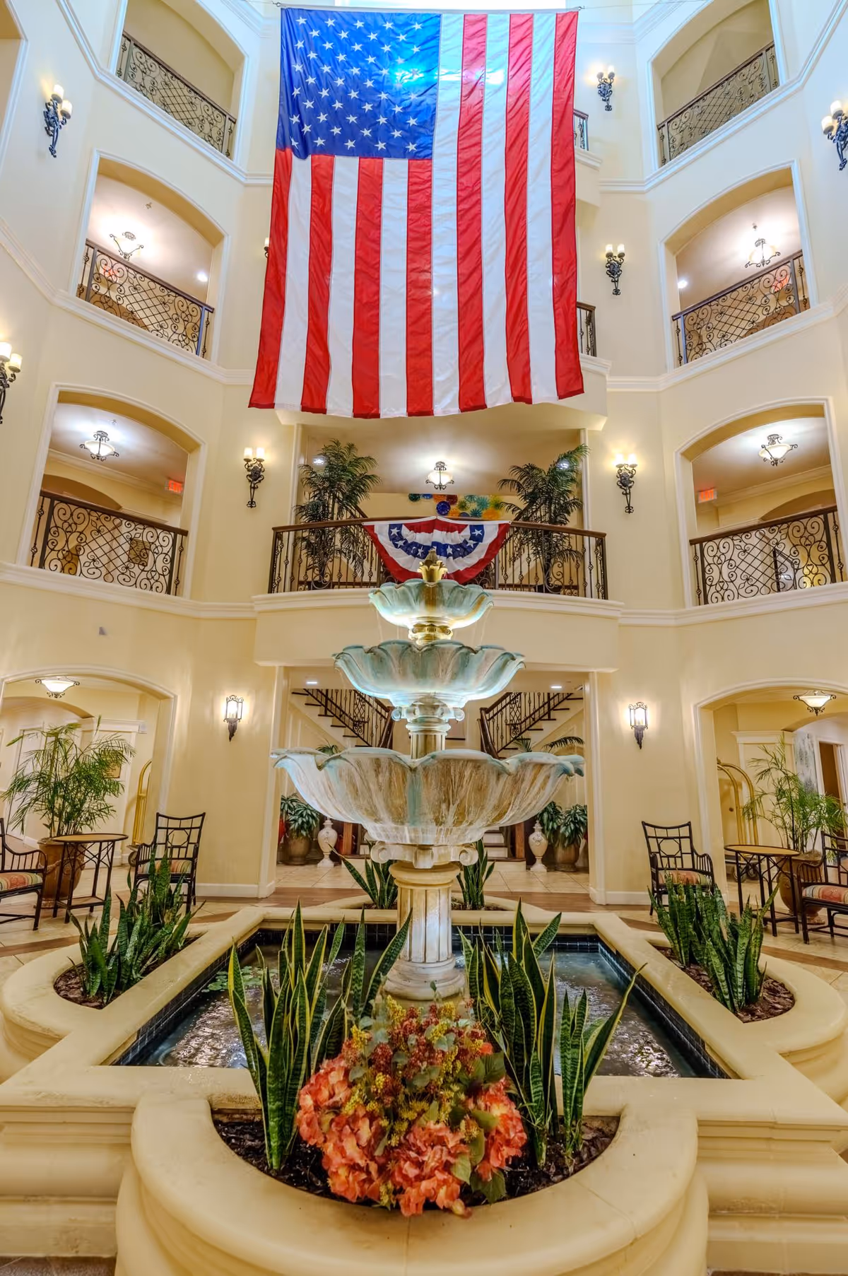Interior view of a multi-story atrium with a large American flag hanging from the upper floor. The space features a tiered water fountain surrounded by plants and flowers in the center, with wrought iron railings on balconies overlooking the area. There are chairs and tables along the sides, and warm lighting fixtures on the walls.