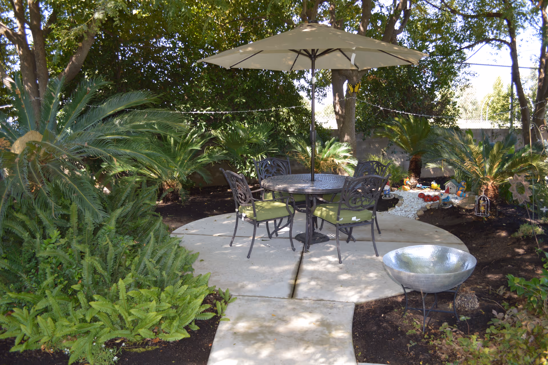 Outdoor garden patio with a round table, umbrella and four chairs surrounded by lush plants and decorative landscaping.