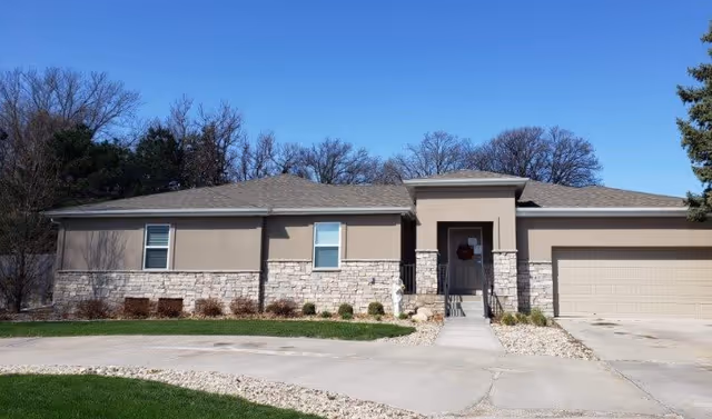Single-story building with beige and stone exterior, a front entrance with a small porch and steps, a two-car garage on the right, and leafless trees in the background under a clear blue sky.