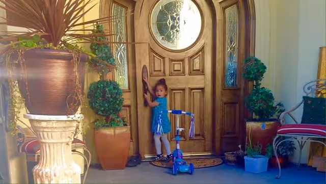 A young child in a blue dress standing at a wooden front door with decorative glass panels, holding the door handle. The entrance area has several potted plants and a blue scooter nearby. There is a metal chair with cushions on the right side.