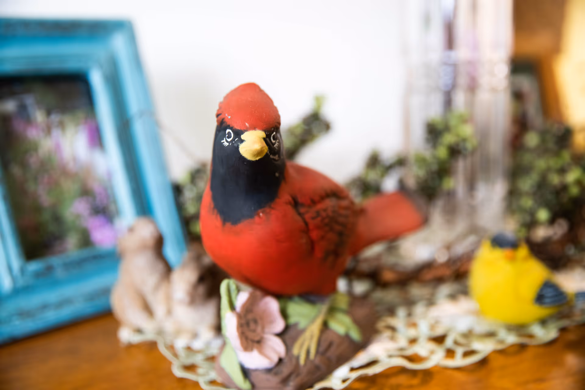 A close-up of a decorative red cardinal bird figurine perched on a floral base, with a blurred yellow bird figurine, a blue picture frame, and other decorative items in the background on a wooden surface.