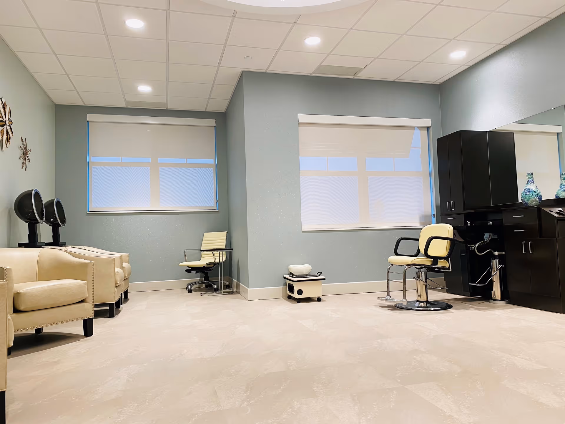 A clean and modern salon area with light beige armchairs on the left, two black hair dryer stations mounted on the wall, a cream-colored swivel chair with armrests near a black cabinet with a sink, and two large windows with white blinds. The walls are painted light blue and the floor is a light beige tile.