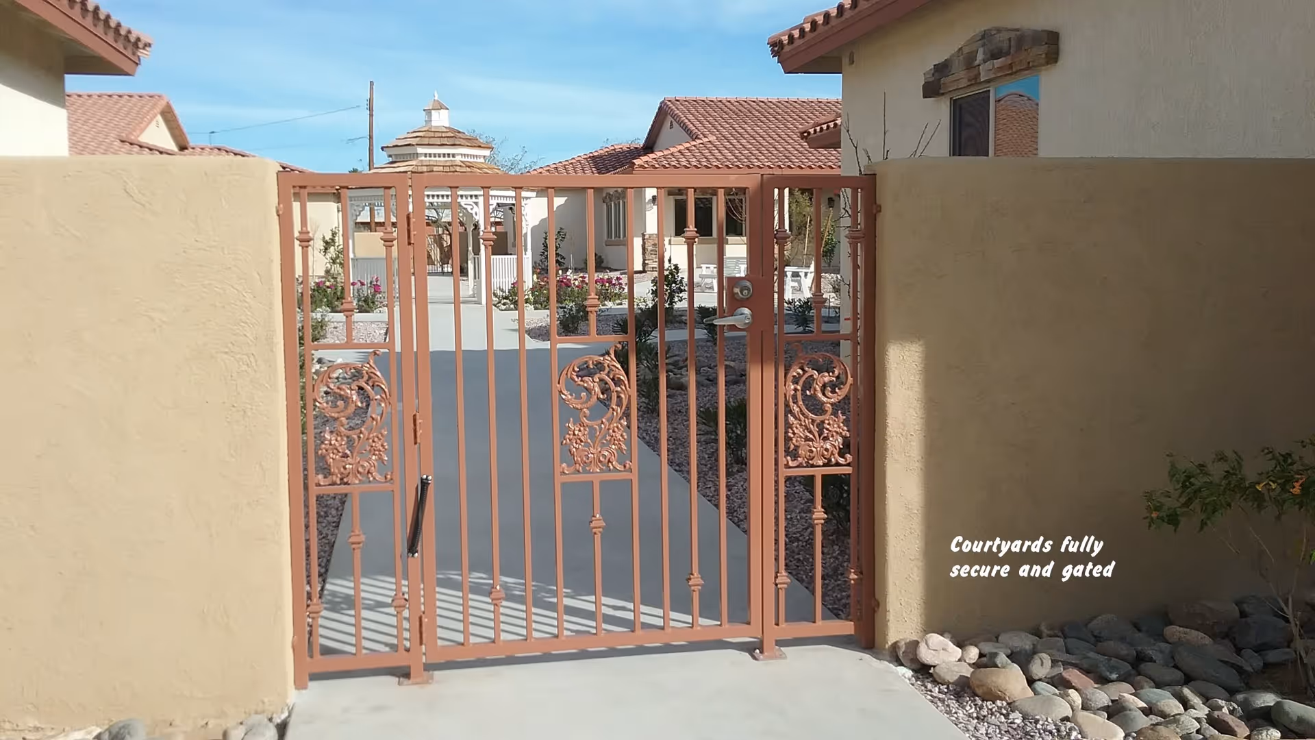 A secure, gated courtyard entrance with a decorative metal gate and beige stucco walls on either side. Beyond the gate, there is a paved walkway leading to buildings with red tile roofs and landscaped areas with plants and rocks. The sky is clear and blue.