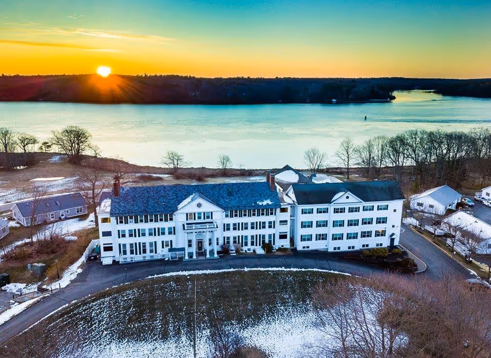 Aerial view of a large white multi-story building with a blue roof, situated near a body of water at sunset. The surrounding area has patches of snow and trees, with a road curving around the building.