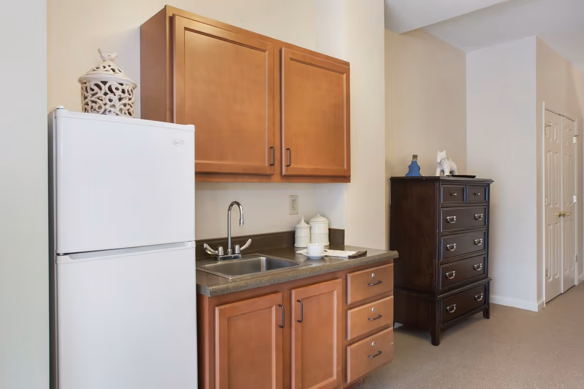 A small kitchenette area with a white refrigerator, wooden cabinets above and below a countertop with a sink. On the countertop are two white canisters and a cup with a saucer. To the right of the kitchenette is a dark wooden chest of drawers with decorative items on top. The walls are light-colored and there is a carpeted floor.