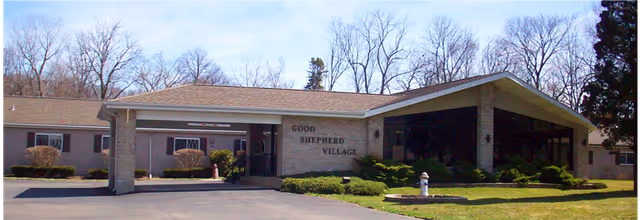 Exterior front view of Good Shepherd Village building with a covered entrance, brick walls, large windows, and surrounding greenery under a clear sky.