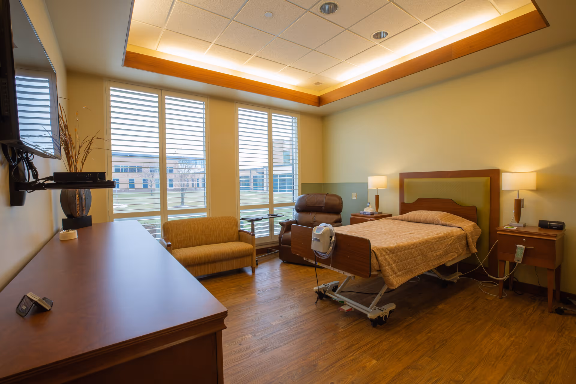 A well-lit senior care bedroom with an adjustable hospital-style bed, recliner, small sofa, TV, and large windows overlooking a courtyard.