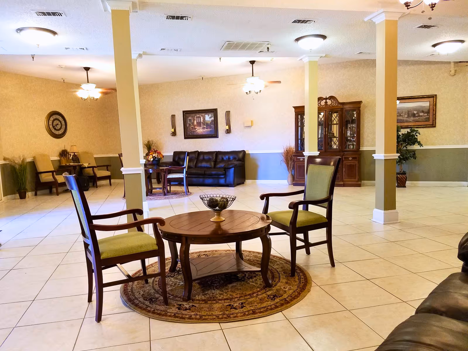 A spacious and well-lit common area with tiled floors, beige walls, and white pillars. The room features a round wooden table with two wooden chairs with green cushions on a circular rug in the foreground. In the background, there is a black leather couch, a wooden cabinet with glass doors, framed artwork on the walls, and additional seating with armchairs. Ceiling fans and light fixtures are mounted on the ceiling.