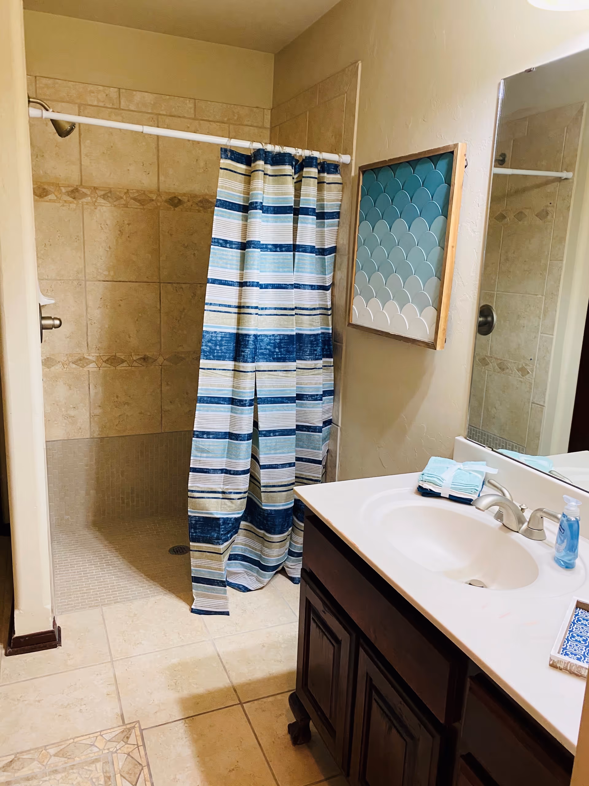 Bathroom with a tiled walk-in shower featuring a striped blue, white, and beige shower curtain. There is a white sink with a silver faucet on a dark wooden vanity. A stack of folded towels and a bottle of blue liquid soap are on the countertop. A decorative wall art piece with blue and white gradient tiles is mounted on the wall next to a large mirror.