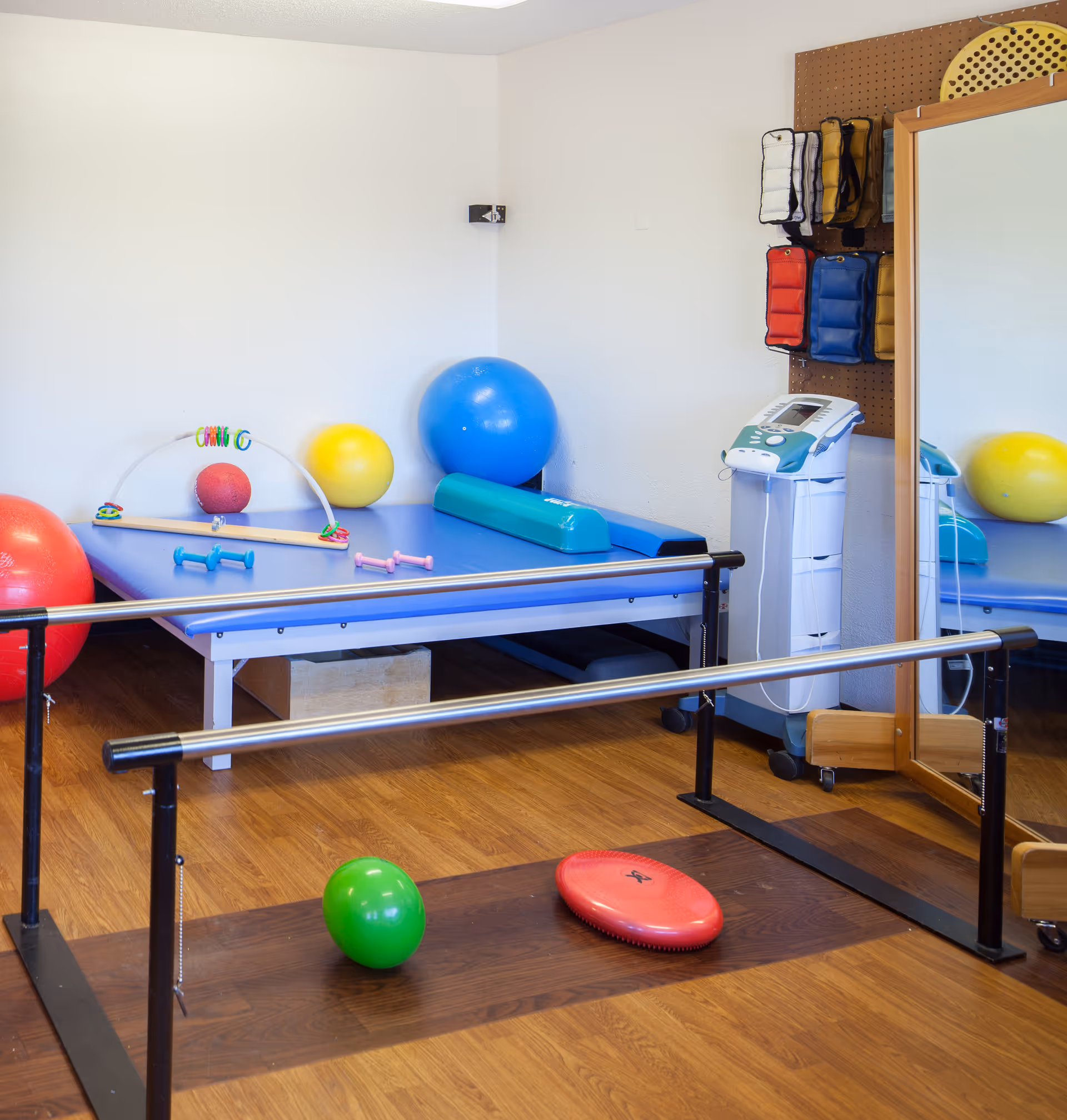 Physical therapy room with parallel bars, exercise balls in various colors, small dumbbells, a large mirror, and therapy equipment on a wooden floor.
