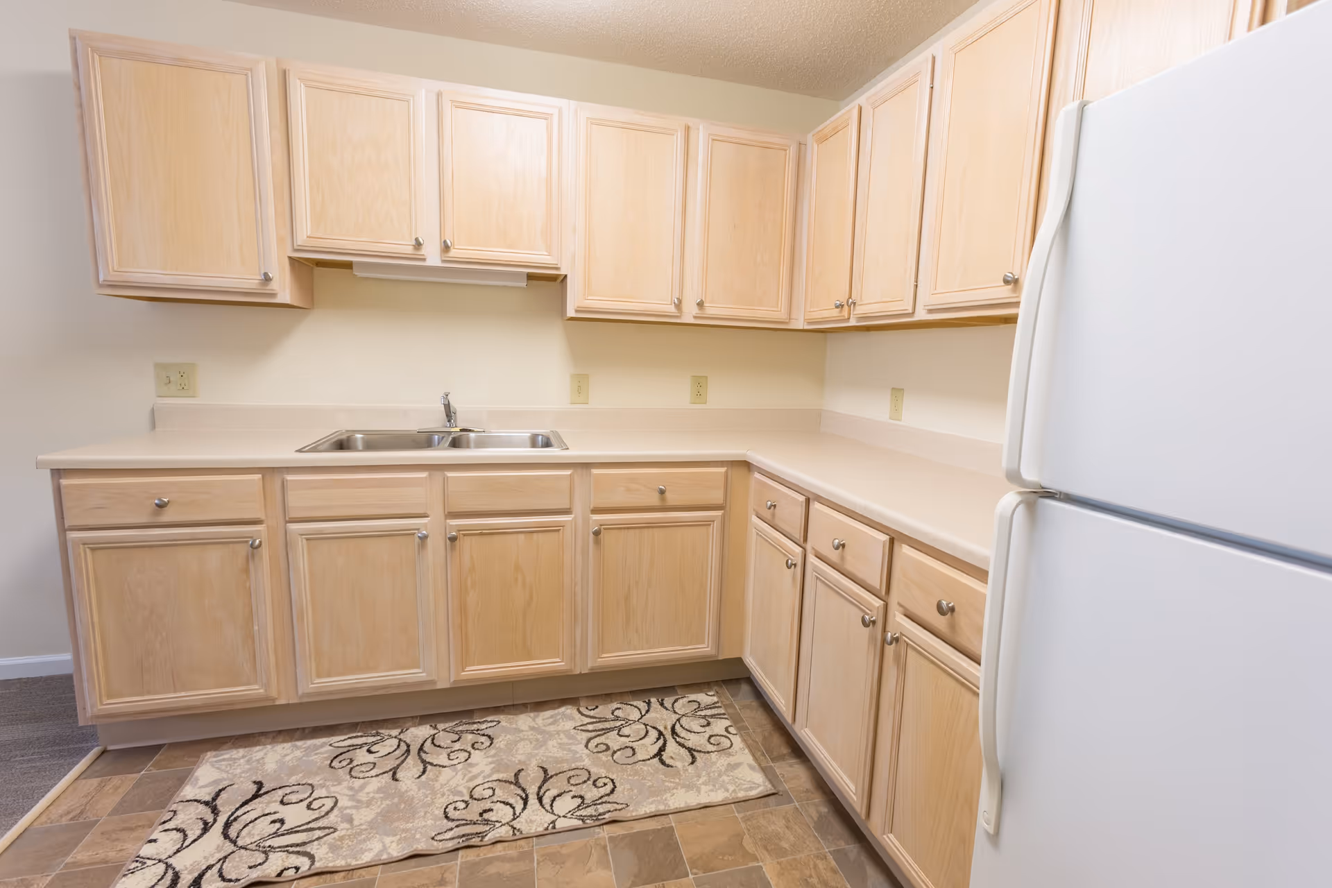 A clean kitchen with light wood cabinets, a double stainless steel sink, beige countertops, a white refrigerator, and a patterned rug on the tiled floor.