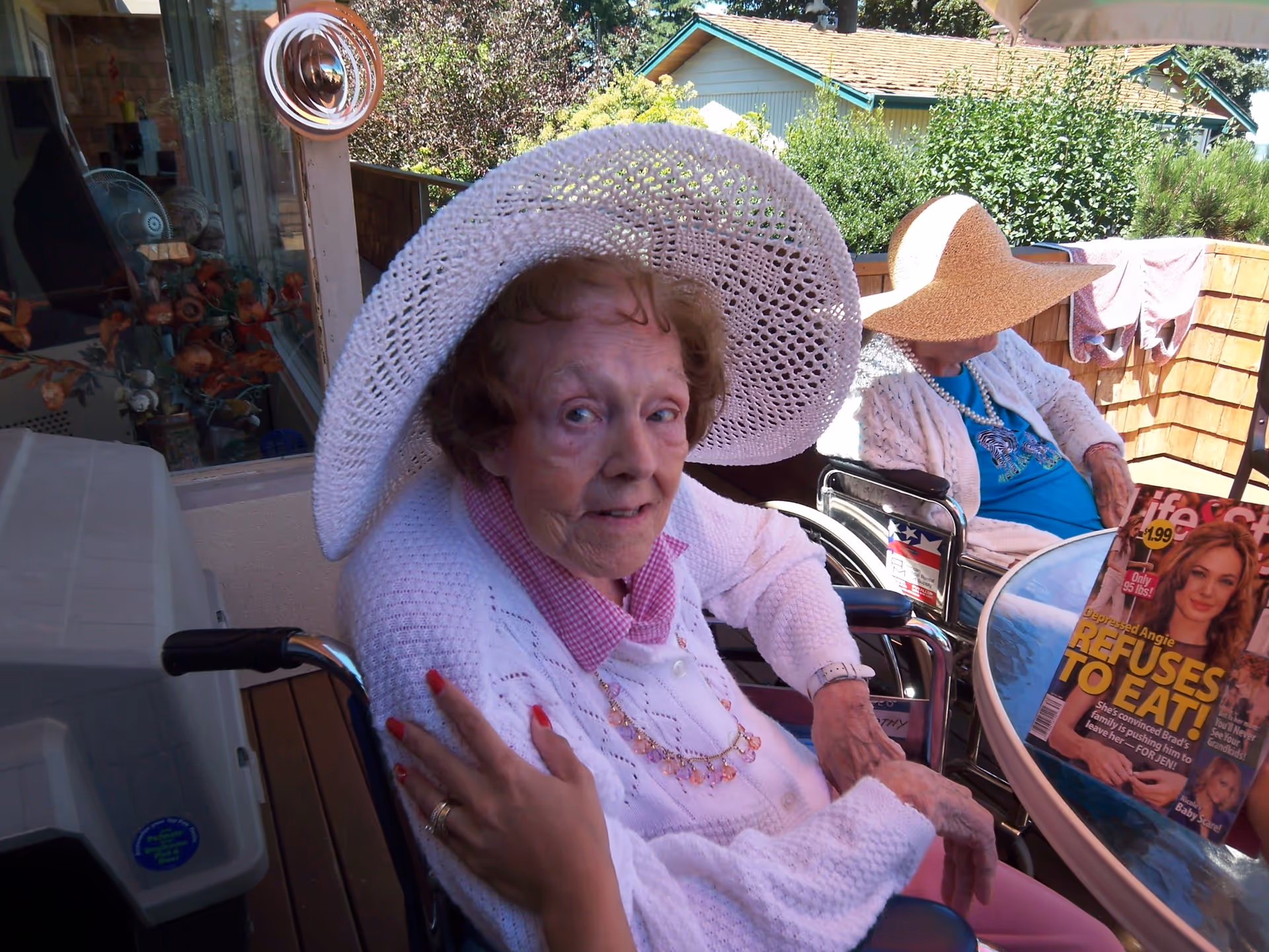 Two elderly women sitting outdoors on a patio, both wearing large sun hats and white sweaters. One woman is looking at the camera while the other is looking down. A magazine with the headline 'REFUSES TO EAT!' is on the glass table in front of them. There is greenery and a house visible in the background.