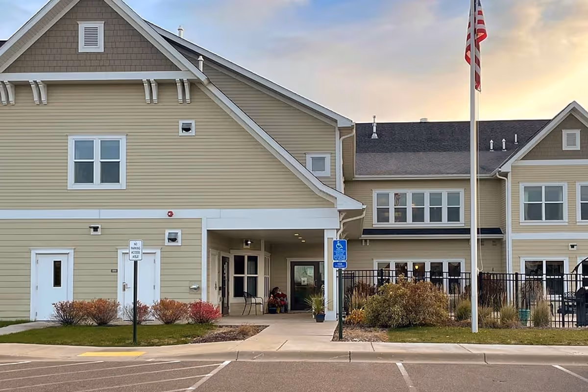 Exterior view of Volante Senior Living of Hanover building with beige siding, multiple windows, an entrance with a covered walkway, landscaping with bushes, a flagpole with an American flag, and parking spaces including a handicapped spot.