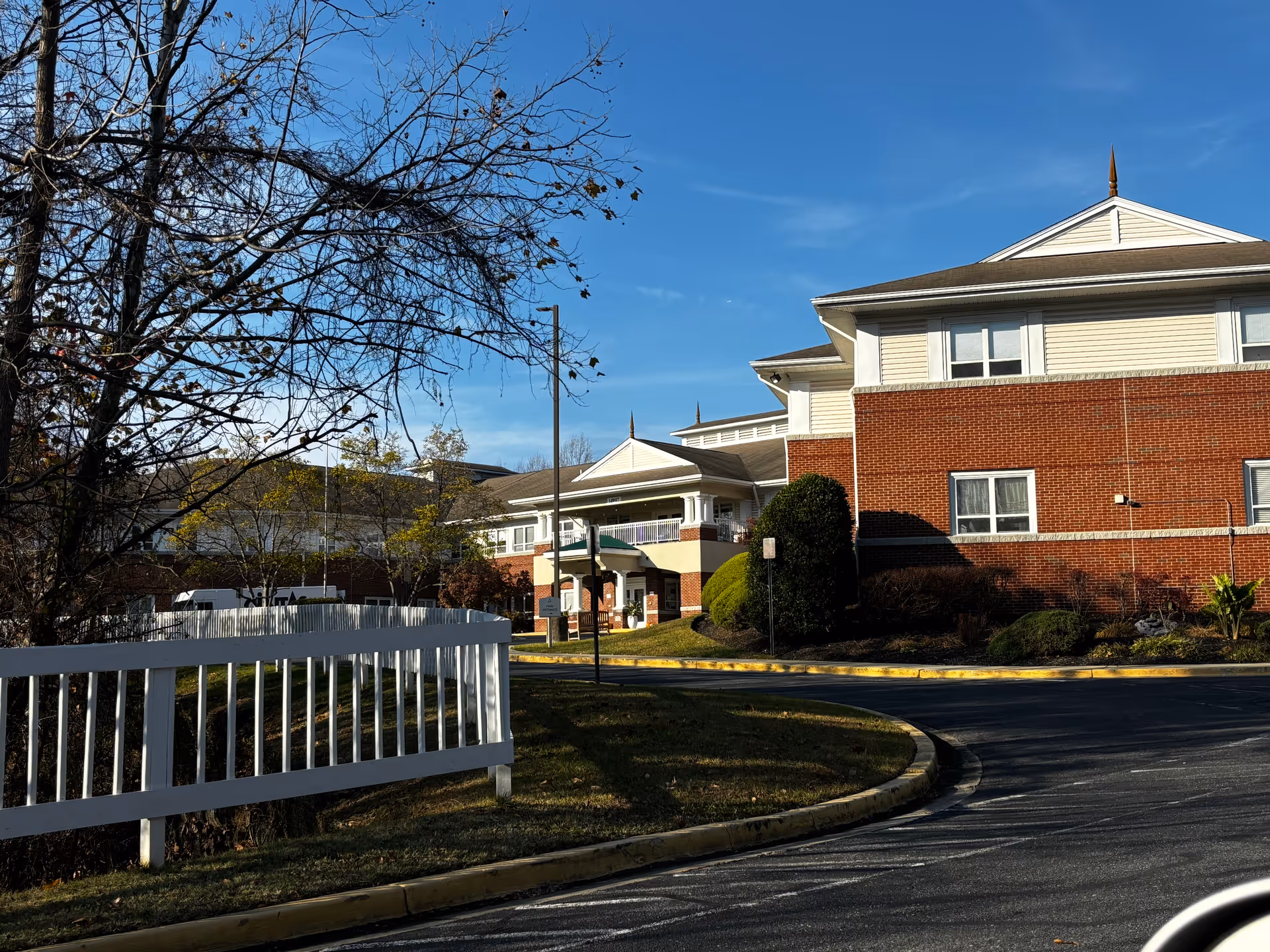 Exterior view of a senior living facility building with red brick and beige siding, a white fence in the foreground, some trees with sparse leaves, and a curved driveway leading to the entrance under a covered porch. The sky is clear and blue.