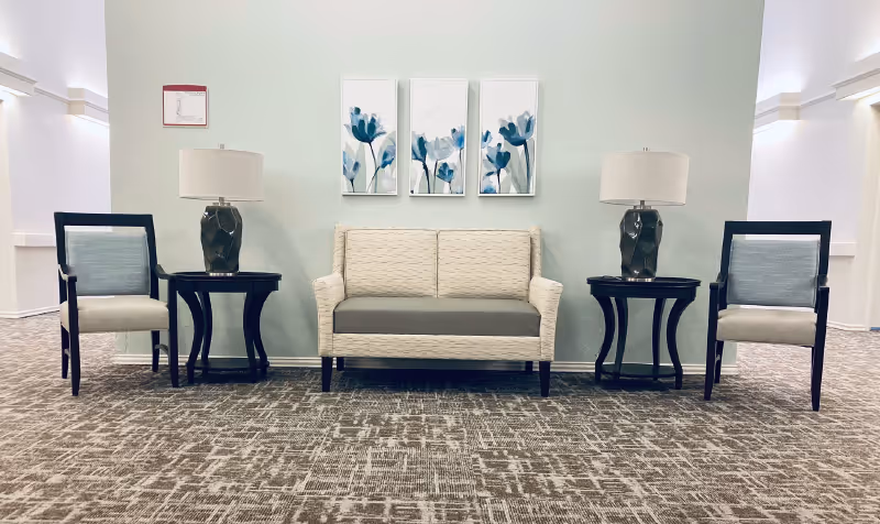 Seating area with a small loveseat flanked by two side tables and lamps and a chair on each side under three floral wall prints.
