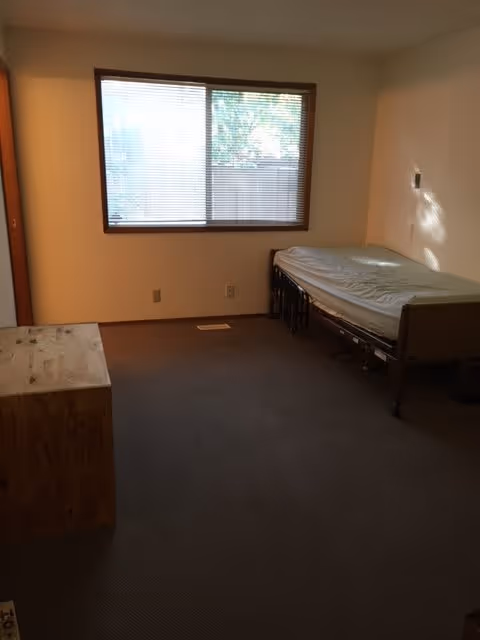 A simple bedroom with a single bed against the right wall, a wooden chest or dresser on the left, a large window with blinds letting in natural light, and beige walls and carpeted floor.