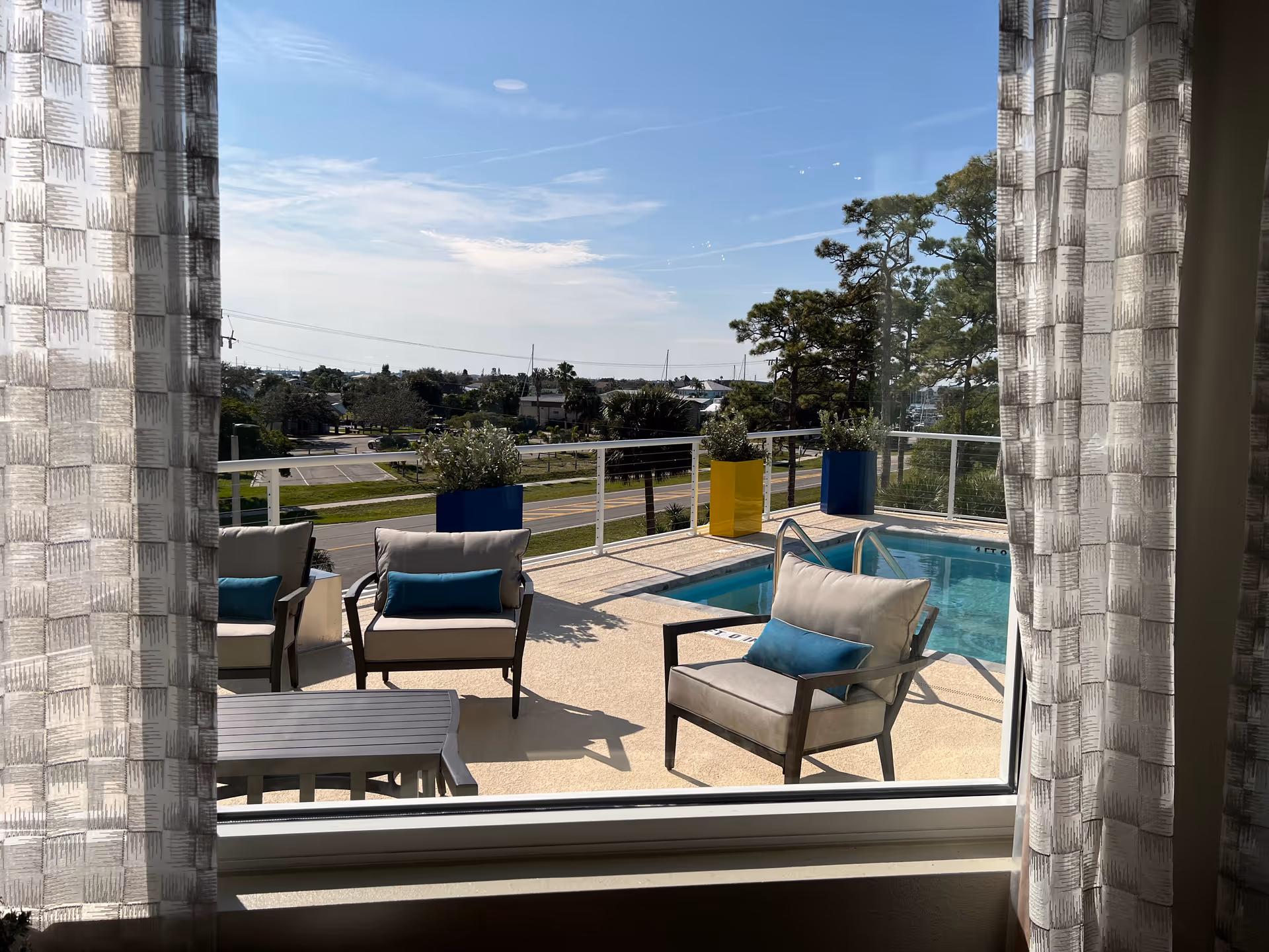 View through a window with patterned curtains of an outdoor patio area featuring cushioned chairs with blue pillows, a small table, potted plants in blue and yellow planters, and a swimming pool. Trees and a road are visible in the background under a partly cloudy sky.