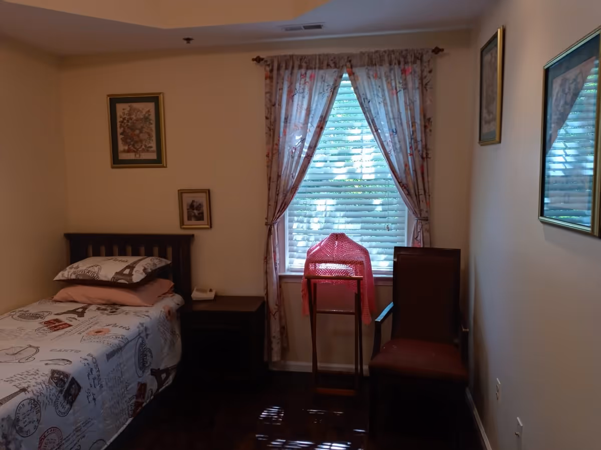 A small bedroom with a single bed covered in a Paris-themed bedspread and pillows. There is a wooden nightstand next to the bed with a telephone on it. A window with floral curtains and blinds is in the center of the wall. In front of the window, there is a wooden stand with a pink knitted cover and a wooden chair with a red cushion. The walls are decorated with framed pictures.