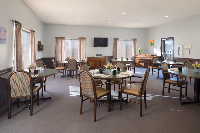 A bright dining room with several round tables and cushioned chairs arranged neatly. Each table has place settings and small floral centerpieces. The room has large windows with curtains, a wall-mounted TV, a wooden sideboard, and a piano near the back wall. The walls are painted light with a darker wainscoting, and the floor is carpeted.