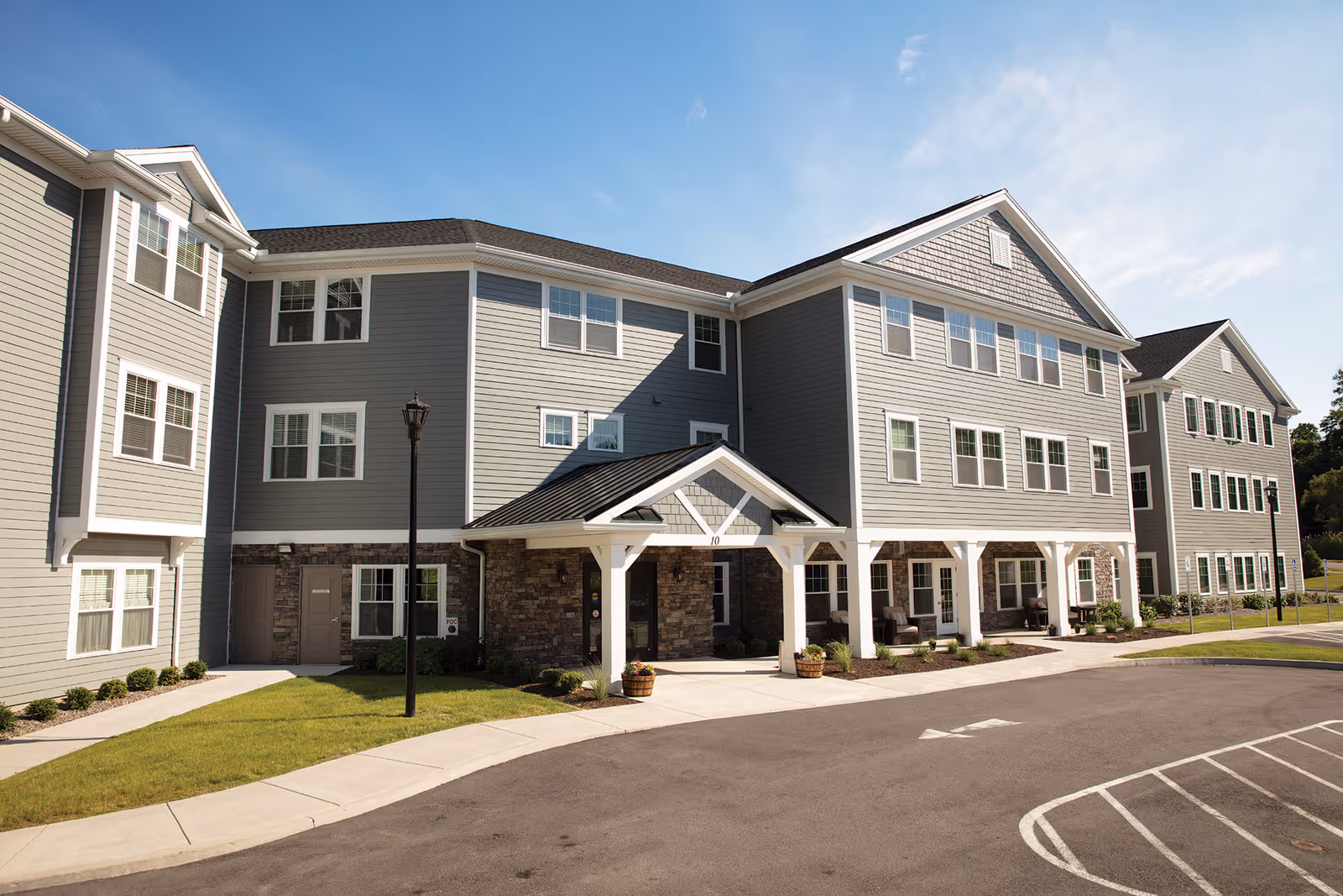 Exterior view of The Woodlands Senior Apartments building with gray siding, multiple windows, a covered entrance, and a parking area in front under a clear blue sky.