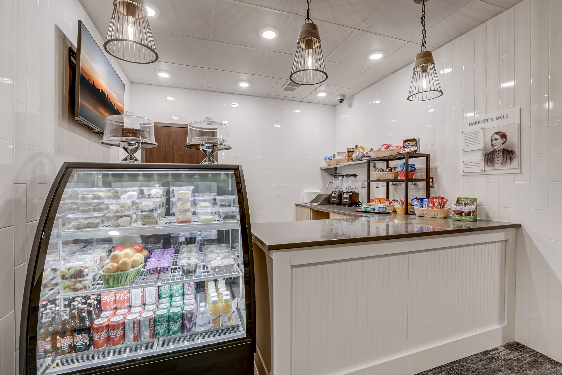 Interior view of a small deli counter area with a glass display case filled with packaged food items, fruits, and beverages. Behind the counter are shelves stocked with snacks and two blenders. The walls are white tiled, and three pendant lights hang from the ceiling. A sign on the wall reads 'Harriet's Deli.'