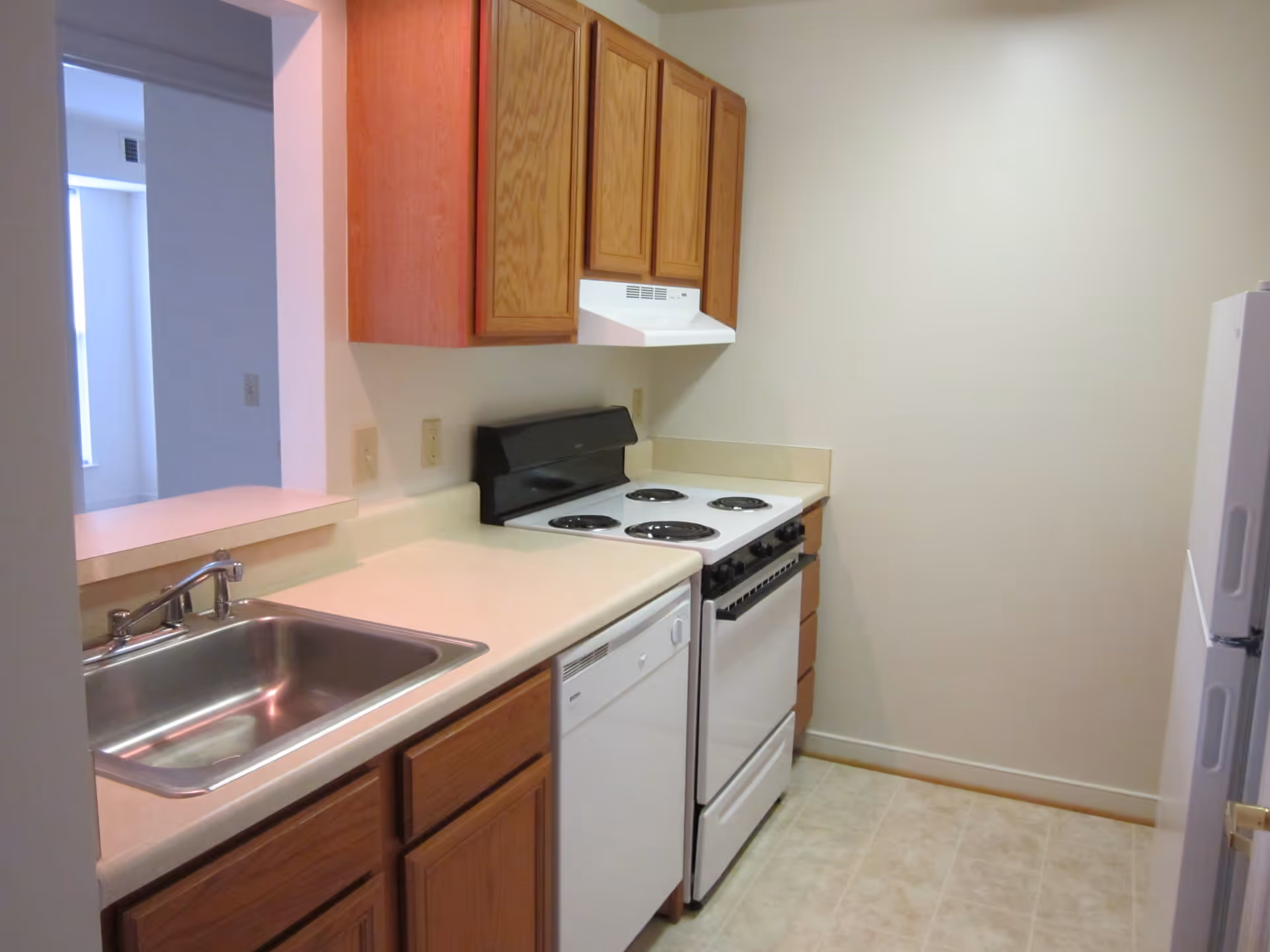 Small galley kitchen with a stainless sink, dishwasher, electric stove, wooden cabinets and a refrigerator.